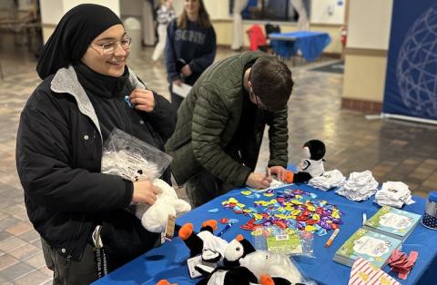 Two students stand in front of a table decorated with black and white penguin plushies, stickers, white socks, and heart-shaped pins. This is for Winter Weekend on campus, where students could make their own winter survival kits for the cold weather.