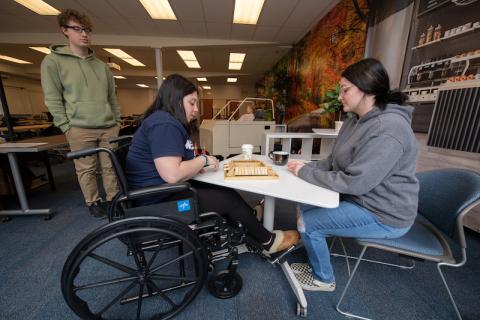 Occupational therapy students. Two sit across from each other on a white table and play jingo. Another stands by and watches.