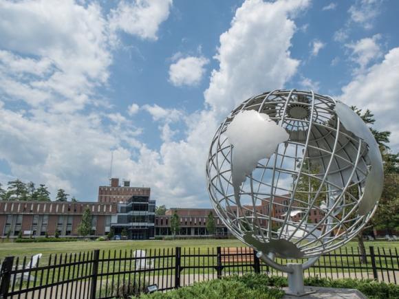 The Westfield State campus globe rests in front of a blue, white-clouded sky. Several brick buildings can be seen in the far background.