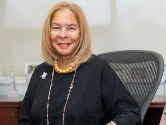 President Linda Thompson at her desk wearing black shirt, gold necklace, and pearls smiling.