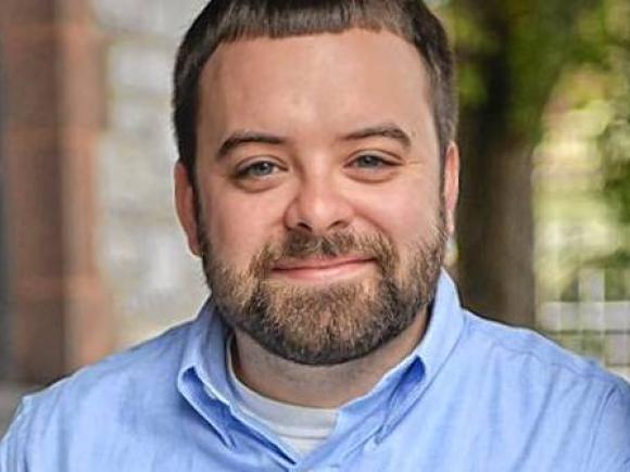 A close-up shot of Joe Courchesne, alumni of Westfield State and teacher at Granby Jr. and Sr. High School. He wears a light blue button-down shirt, and smiles at the camera with blurred out buildings and trees in the background.