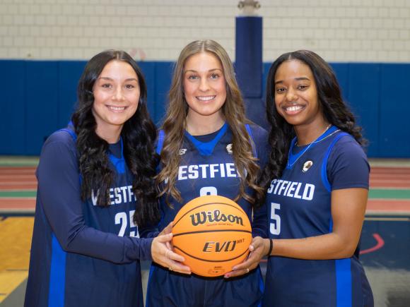 Three women’s basketball players smiling together while holding a basketball.
