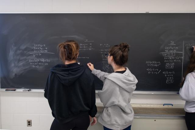 Two students with pony tails and hooded sweatshirts doing math equations on chalkboard