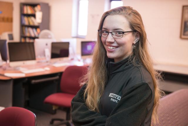 A student smiling in a computer in a lab, with monitors and equipment in the background.