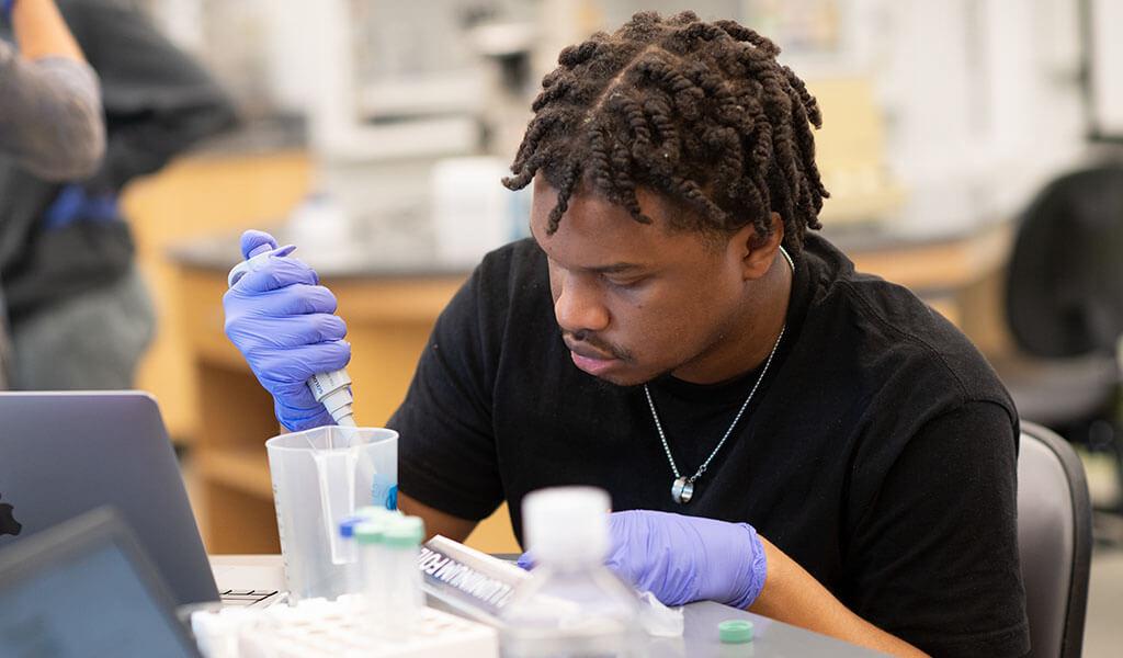 A student wearing rubber gloves conducts an experiment in a laboratory setting. 