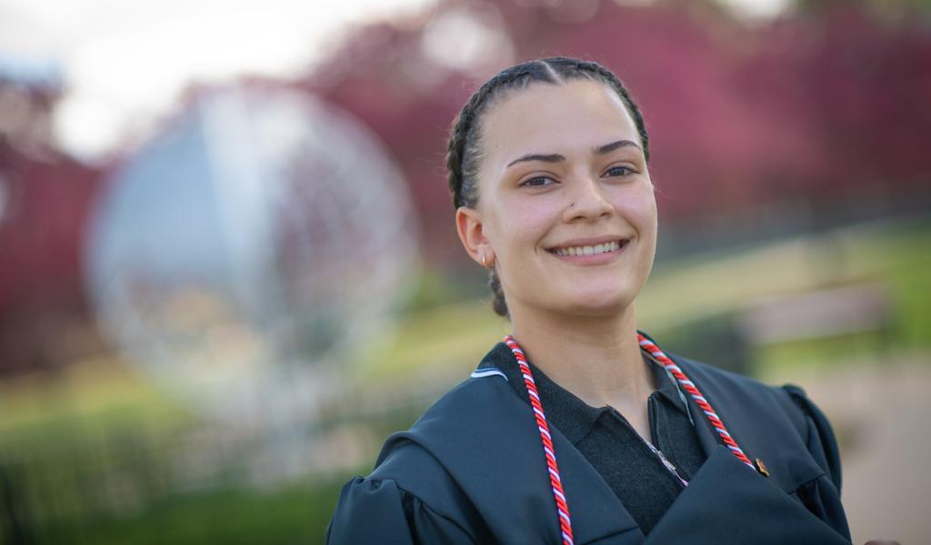 Student in front of the campus globe wearing Commencement gown smiling.