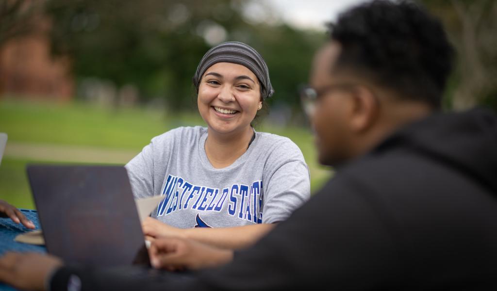 Student smiling outside on campus green with laptop.