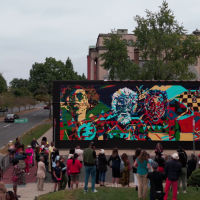 A crowd gathers outside a mural by AfriCOBRA raist and muralist Nelson Stevens in the documentary "Art for the People," directed by Evan Goodchild. Credits to Evan Goodchild and Goodchild Media.